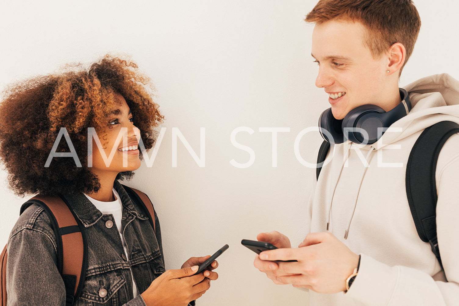 Two classmates standing at wall and looking to each other holding smartphones