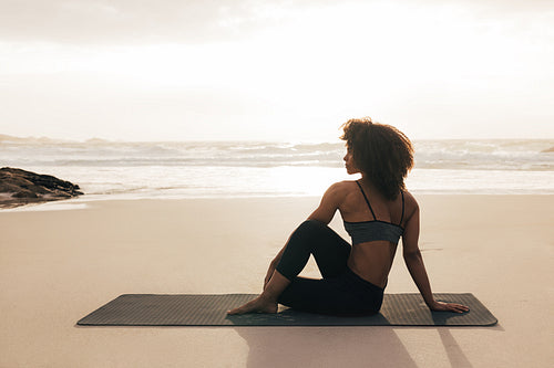 Side view of woman sitting on mat. Female meditating on seashore at sunset.