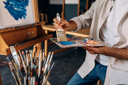 Cropped shot of artist sitting in front of an easel and dipping a large paintbrush into a colorful palette