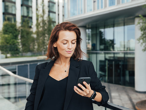 Middle-aged businesswoman looking at her mobile phone while leaning a railing against an office building