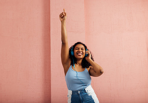 Plus size woman wearing blue headphones raise hand up and dancing against pink wall