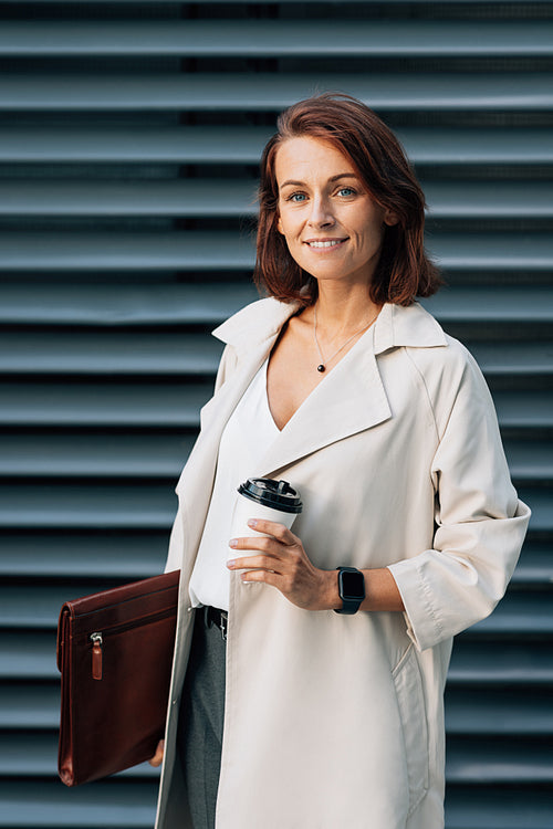 Middle-aged businesswoman holding a coffee to go cup and leather folder while standing outdoors