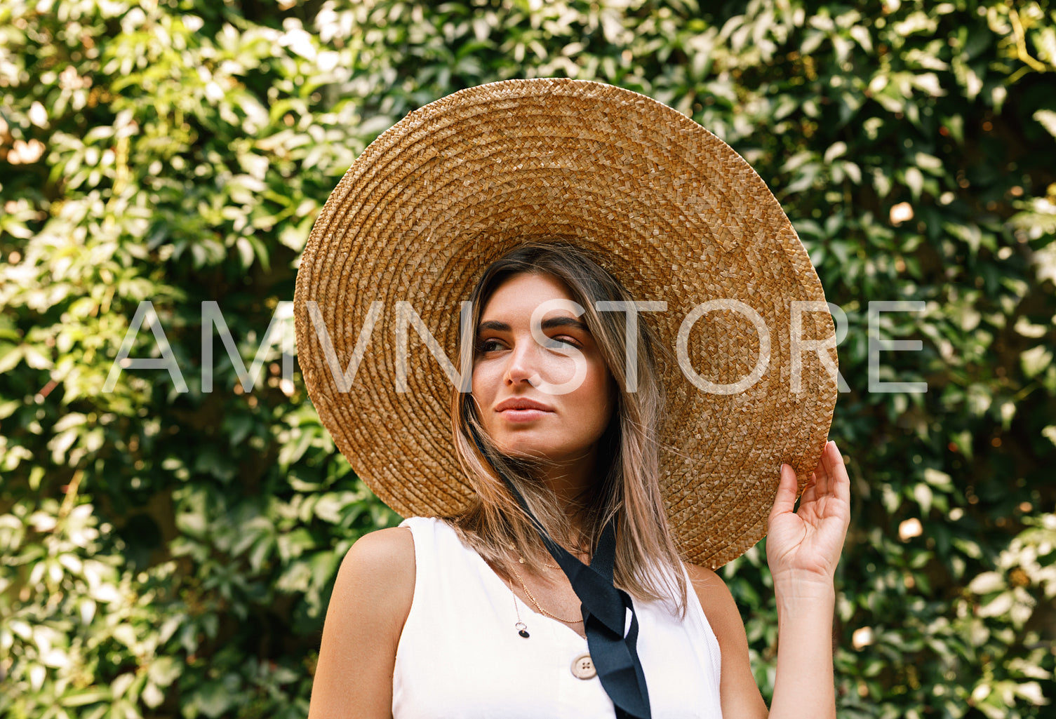 Young woman wearing a straw hat standing outdoors in front of a wall with green leaves	