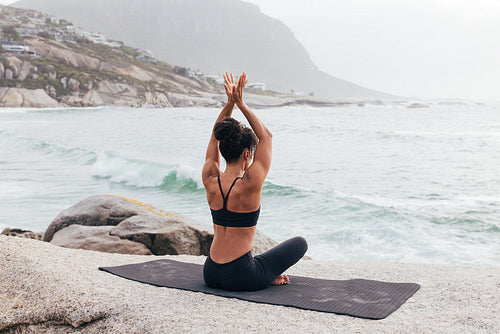 Rear view of fit female sitting on mat with raised hands looking at ocean and waves. Woman relaxing in yoga pose.