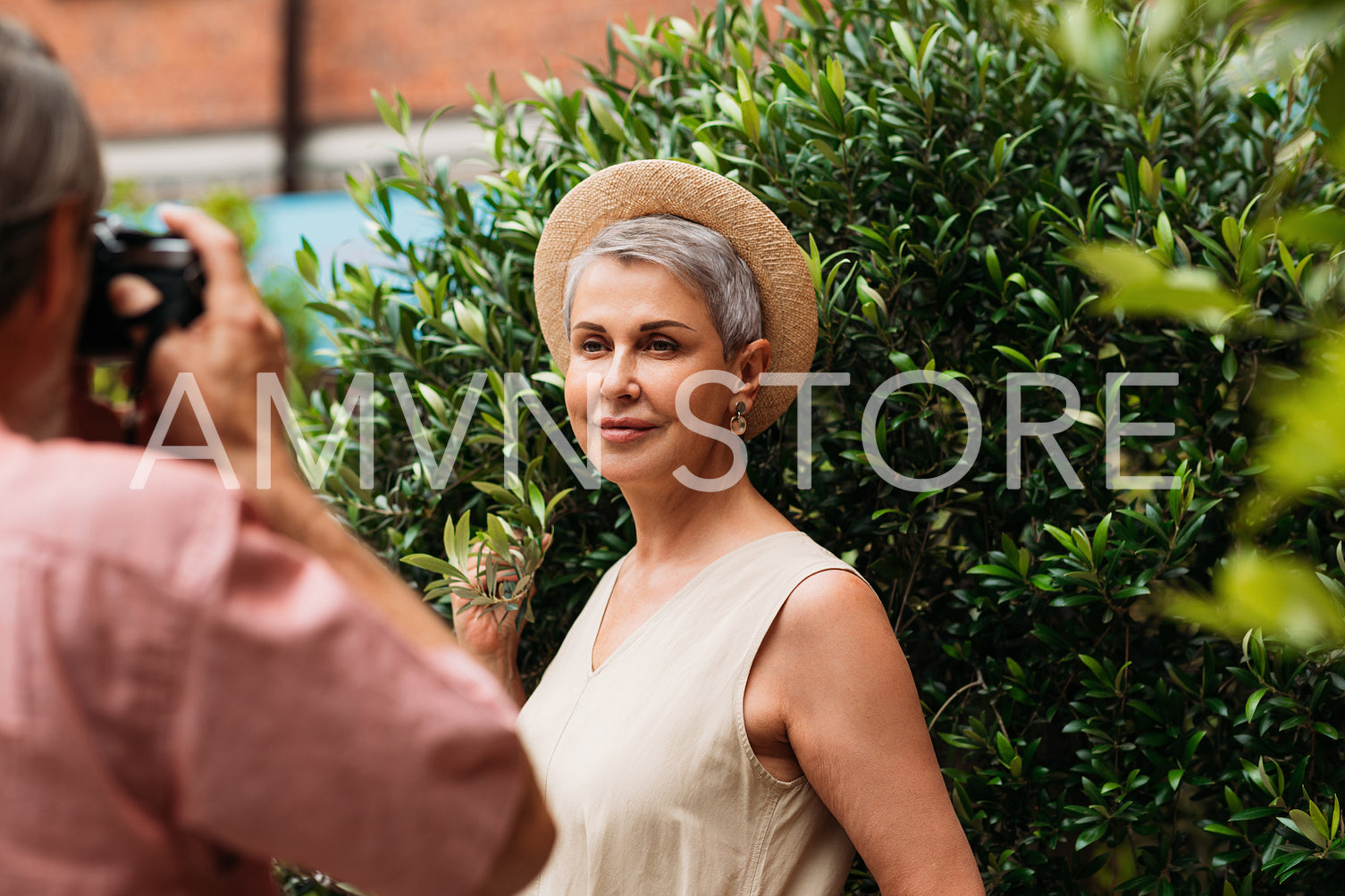 Aged woman with short grey hair and a small straw hat posing against bush while her husband photographs her