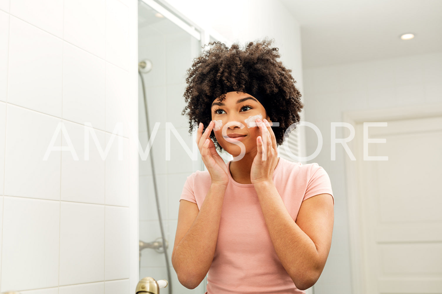 Mixed race woman in bathroom looking into the mirror and applying moisturizer on her face	