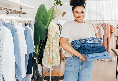 Smiling female fashion store worker holding a pile of jeans and looking at camera