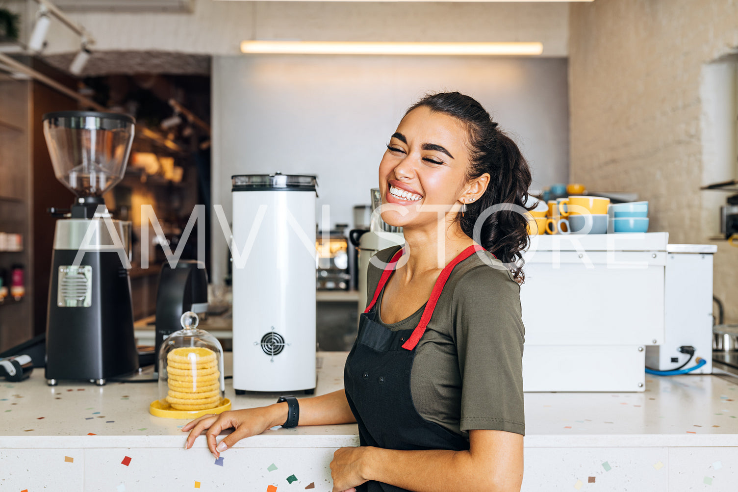 Smiling barista standing at counter in coffee shop with closed eyes. Portrait of a happy waitress wearing apron in cafeteria.	