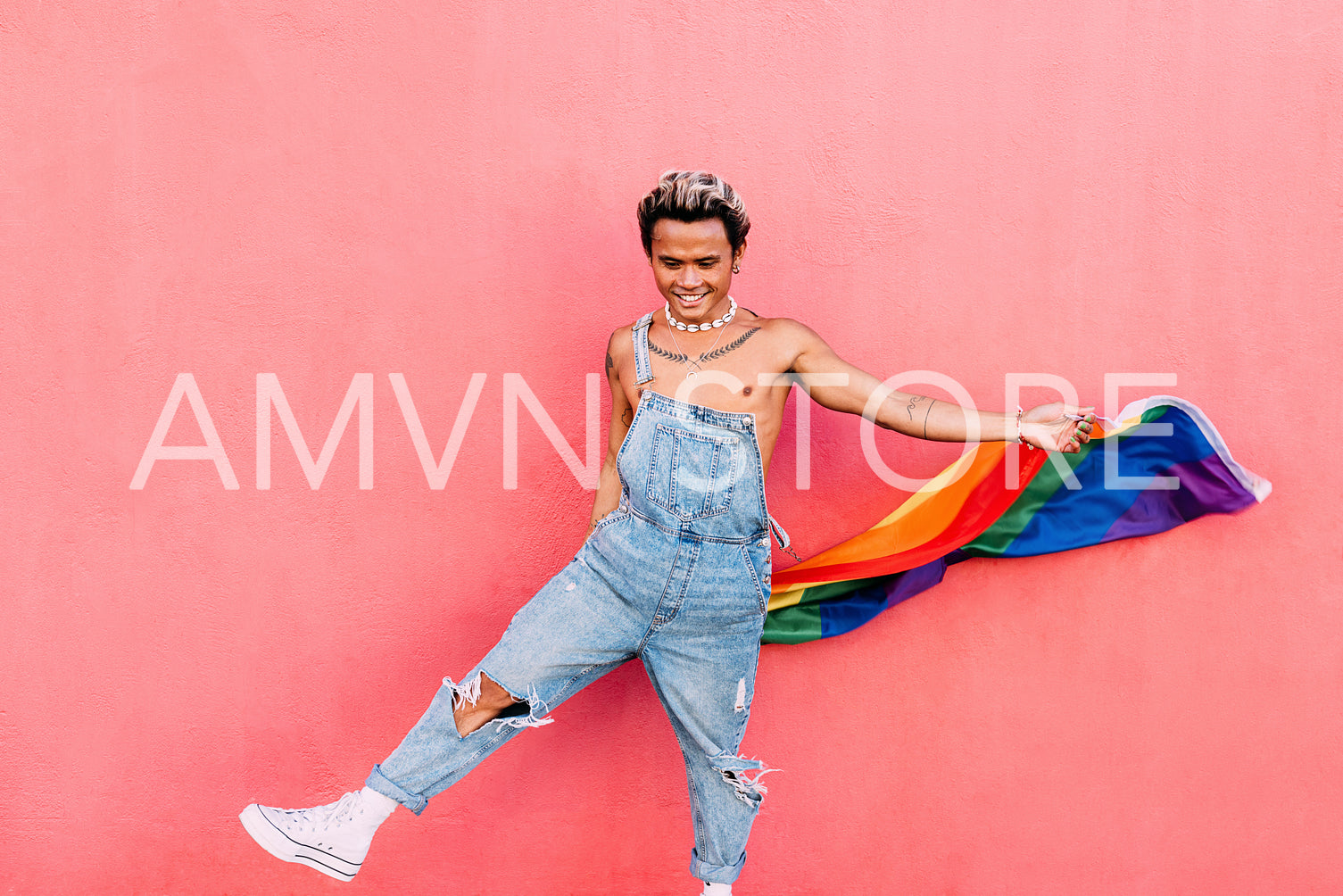 Smiling guy dancing with rainbow LGBT flag against pink wall outdoors