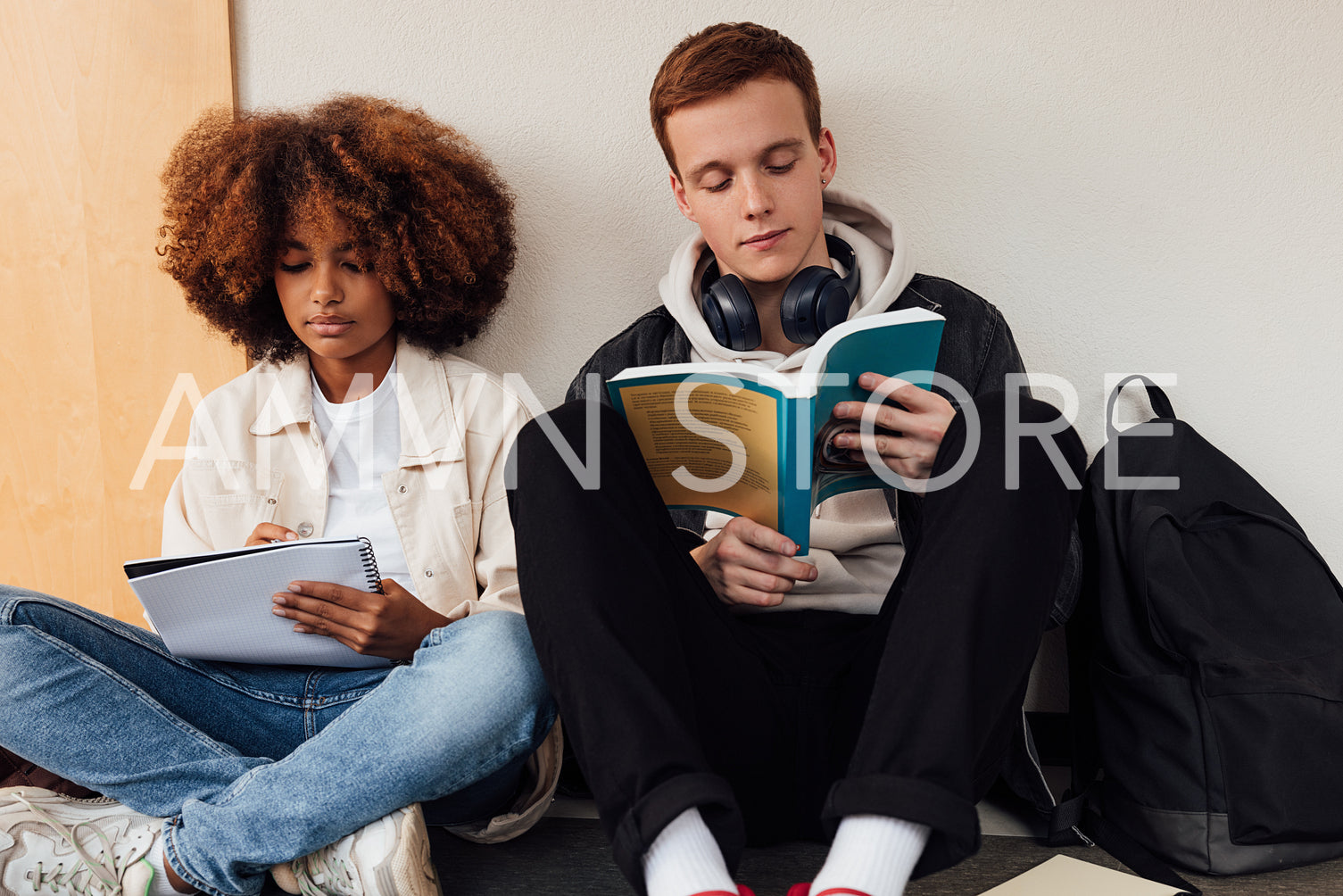 Two university students sitting at wall and reading. Classmates sitting together in corridor with books.