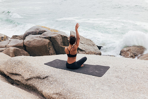 Rear view of fit female raising hand while changing yoga pose. Woman sitting on mat by shore and practicing yoga.