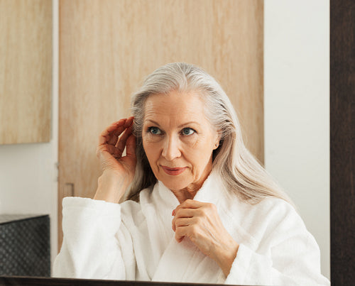 Senior woman adjusting and examines her hair in bathroom mirror in morning