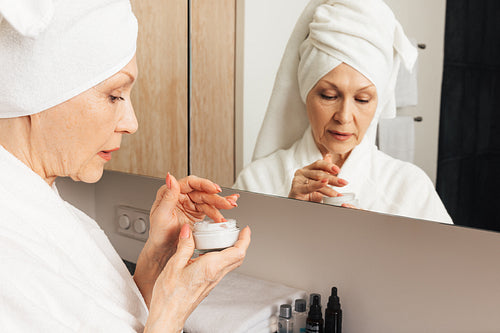 Senior woman with a wrapped towel on a head holding a cream. Aged female preparing to apply a moisturizer in bathroom.