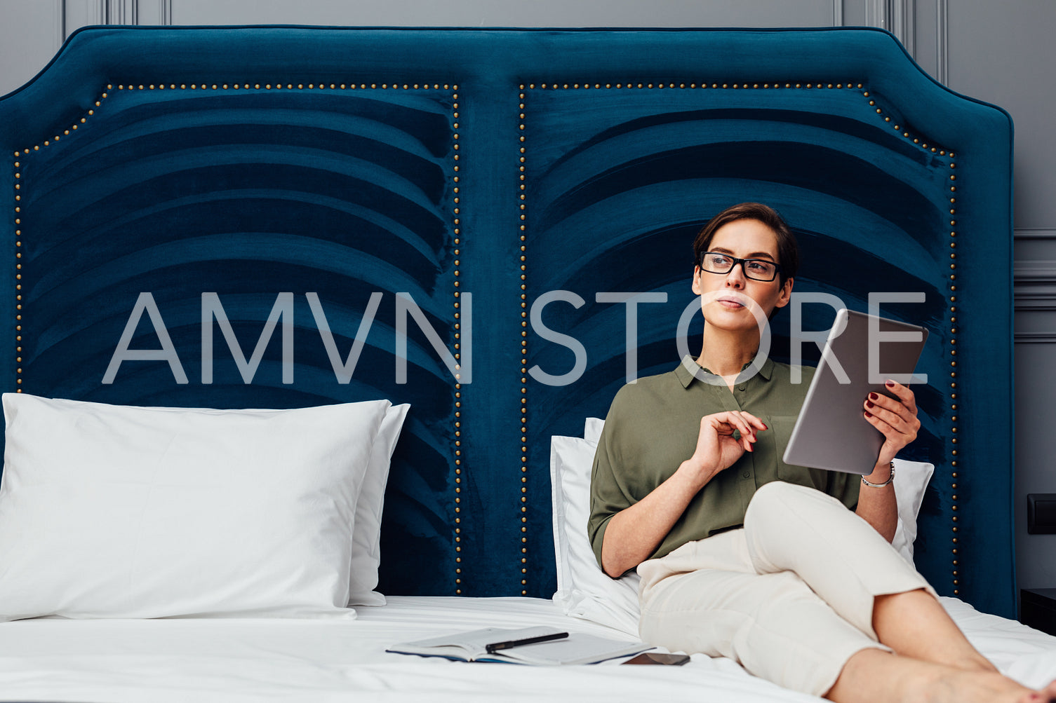 Young woman holding a digital tablet and looking away while lying on a bed in a luxury hotel room	