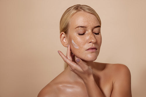 Caucasian woman applying facial moisturizer standing in studio against pastel background