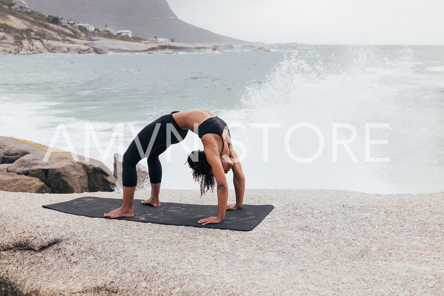 Woman doing Upward Bow pose by ocean with big splashes. Female exercising outdoors at seaside. 