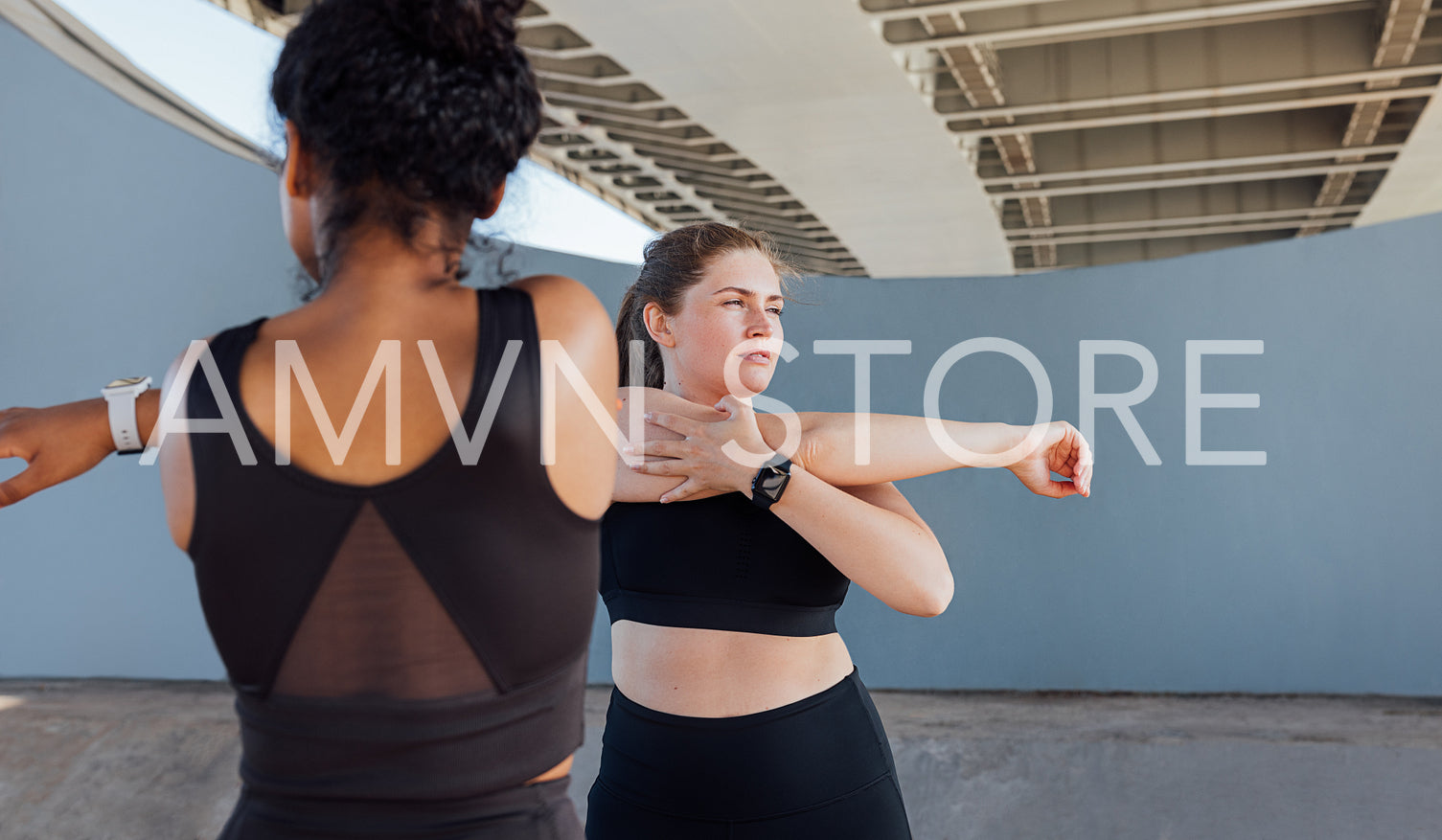 Two plus-size females in black fitness attire warming up. Young women stretch their hands under a bridge.