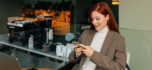 Redhead woman in formal wear sitting in a cafe holding a mobile phone and smiling