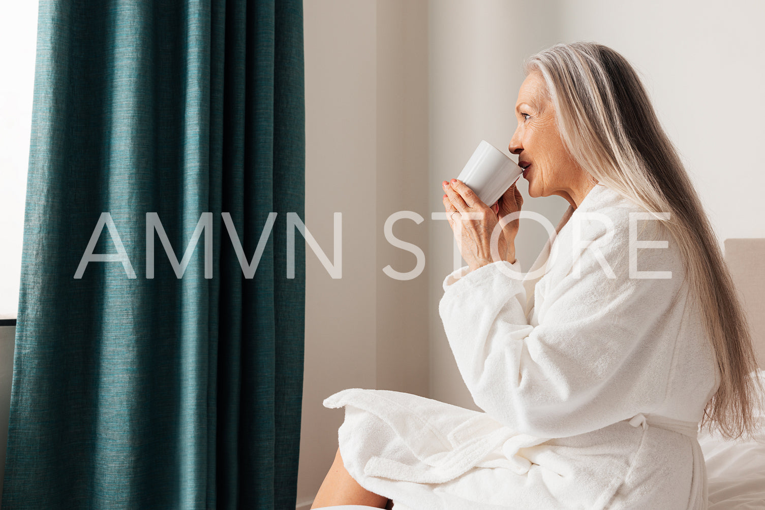 Senior woman drinking coffee in morning in bedroom. Female in bathrobe sitting on a bed looking at window.