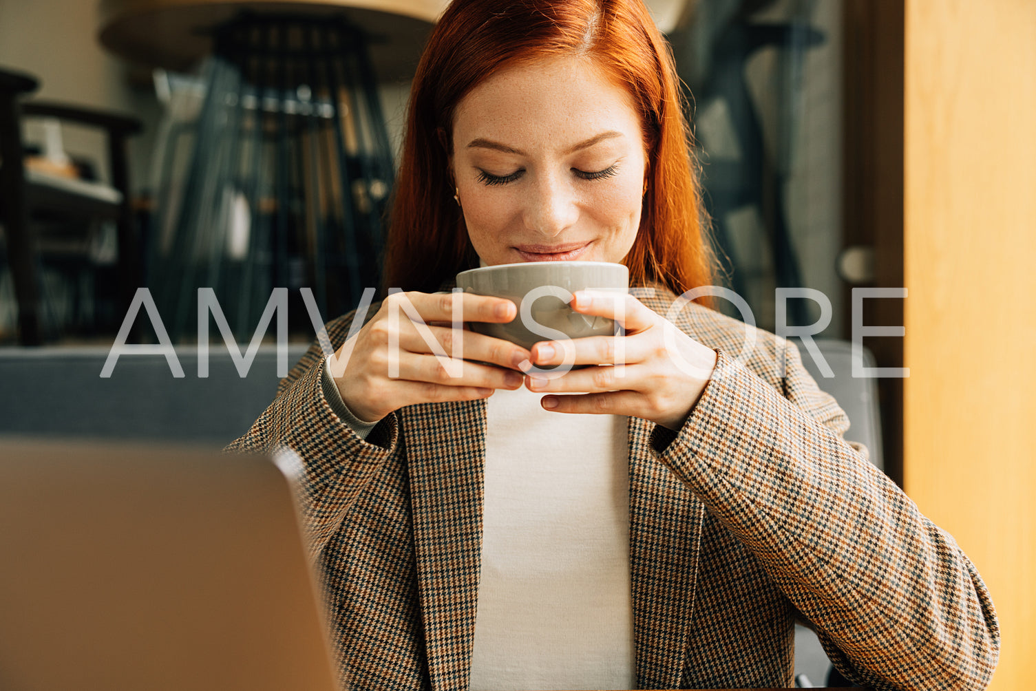 Close-up of a young businesswoman holding a cup and smiling. Female with ginger hair enjoying her coffee in the morning in a cafe.