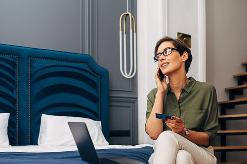 Mid adult woman talking on mobile phone and holding a credit card. Businesswoman making online order while sitting in a luxury apartment.