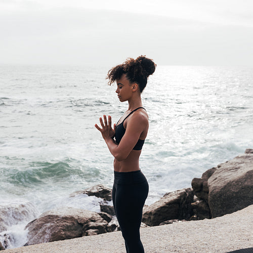 Female with closed eyes and folded hands standing by the ocean at sunset