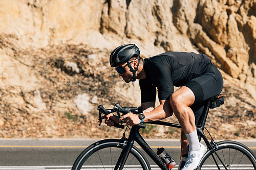 Professional road bike rider in black sportswear. Side view of a young male riding bicycle against a mountain.