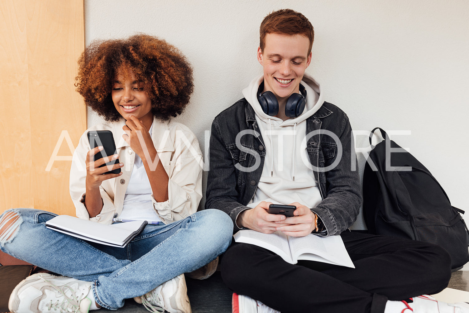 Two smiling classmates sitting at wall and using their smartphones