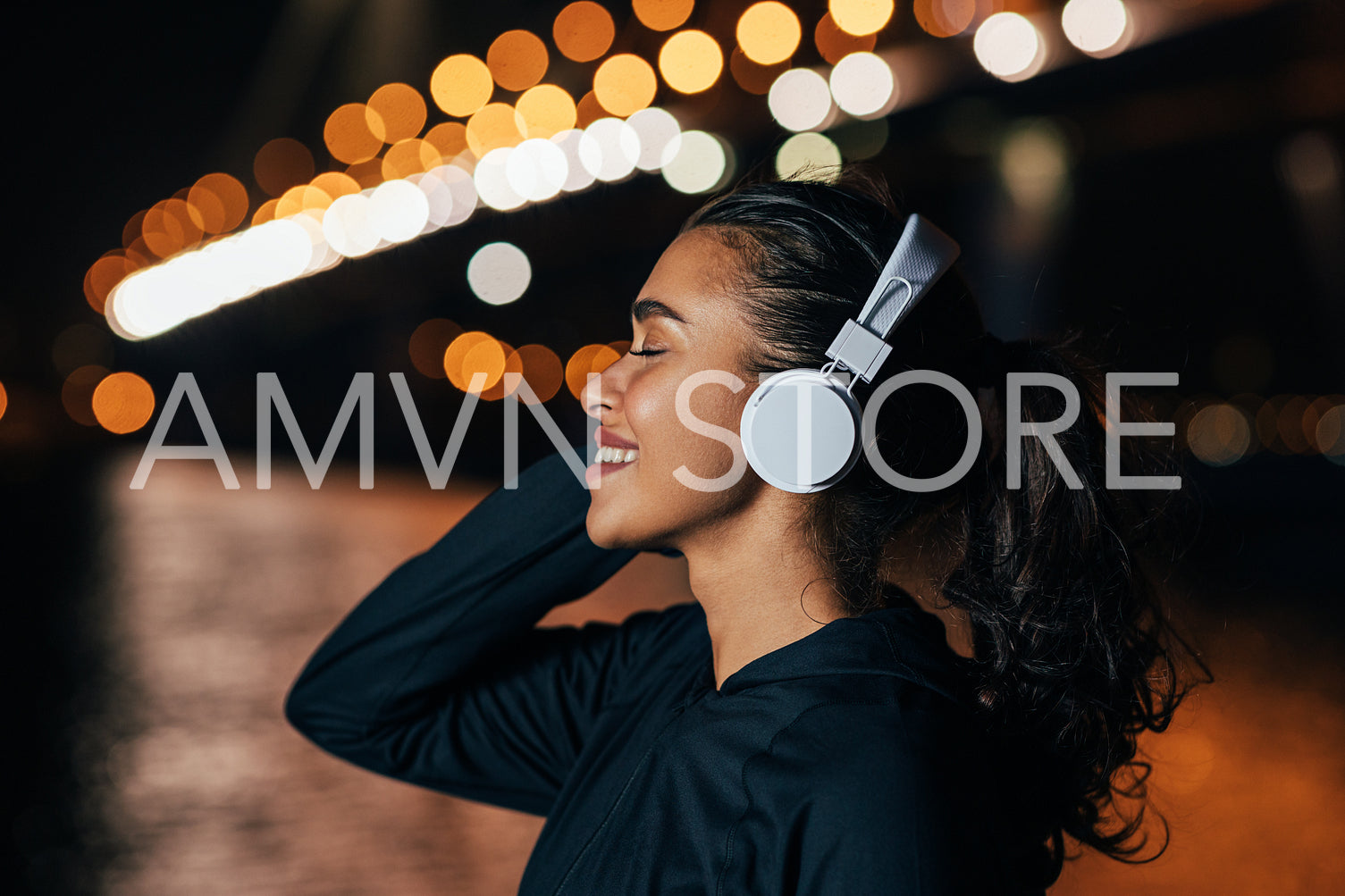 Side view of a fit woman in hooded shirt listening to music against night lights