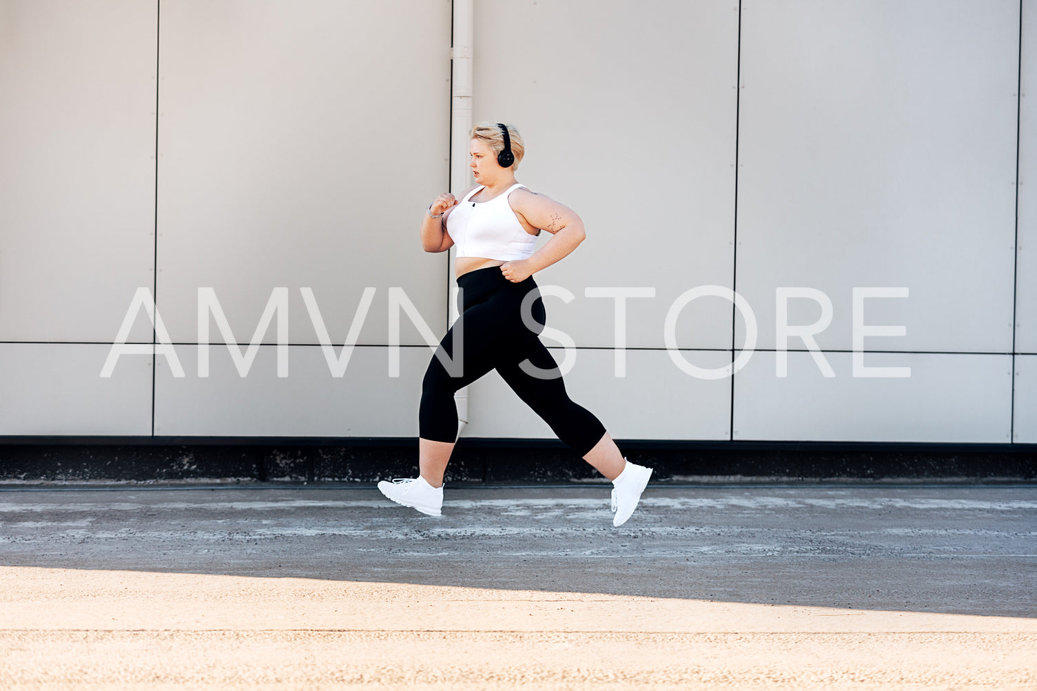 Side view of plus size woman wearing headphones running near a wall. Curvy female in sports clothes jogging outdoors.	