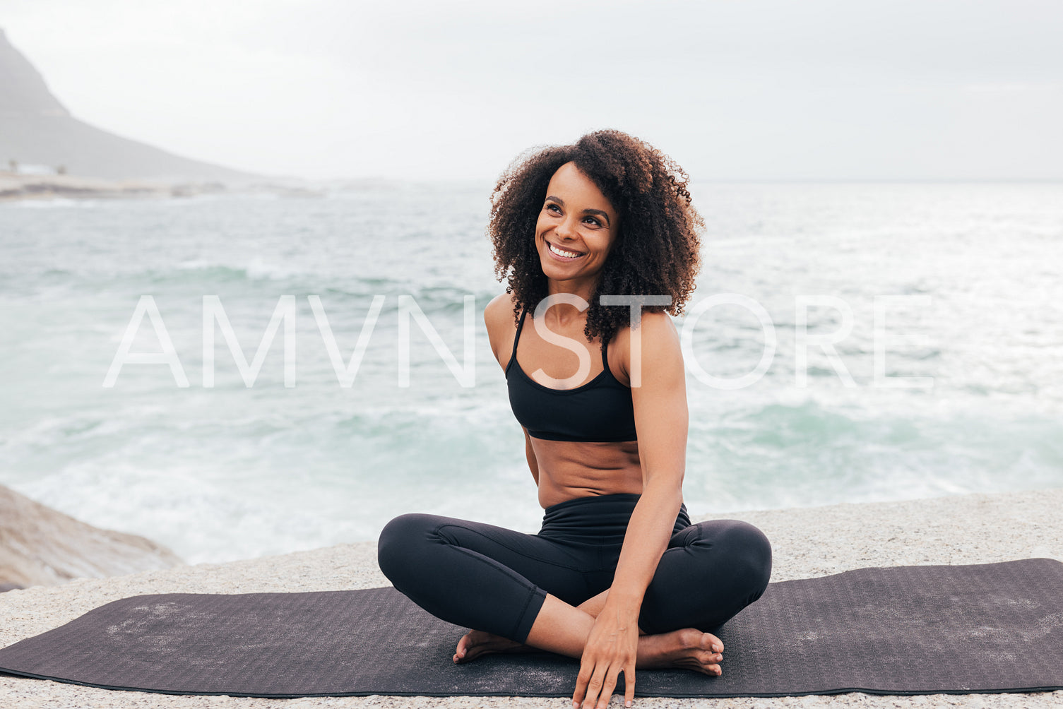 Portrait of young smiling female in sportswear sitting on yoga mat by ocean