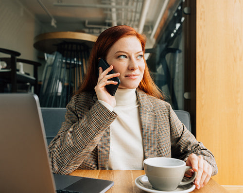 Beautiful businesswoman with ginger hair talking on smartphone and looking at window at a cafe