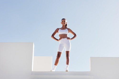 Slim smiling woman in white fitness attire posing on a top of stairs case. Full-length fit female with hands on her hips looking at the camera while standing on a top.