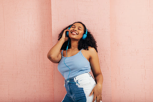 Young woman enjoying music while leaning pink wall. Female wearing blue headphones with closed eyes.