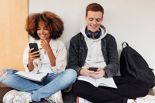 Two smiling classmates sitting at wall and using their smartphones