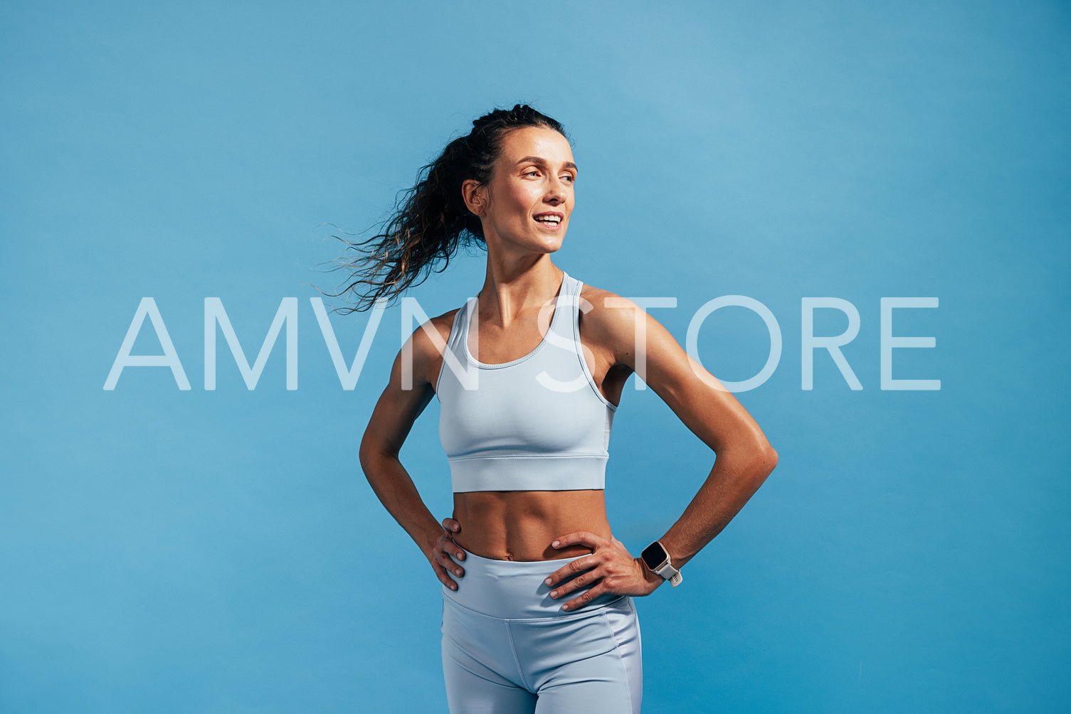 Smiling muscular woman in fitness wear standing on blue background. Female with hands on her hips relaxing after training.