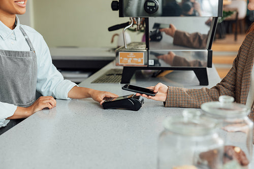 Side view of a barista at counter receiving payment from a customer in a cafe. Unrecognizable female paying by mobile phone using NFC at the counter.