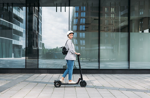 Side view of young smiling businesswoman with cycling helmet on her head driving an electrical push scooter