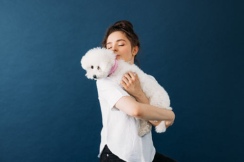 Young woman holding her little white dog in studio over blue bac