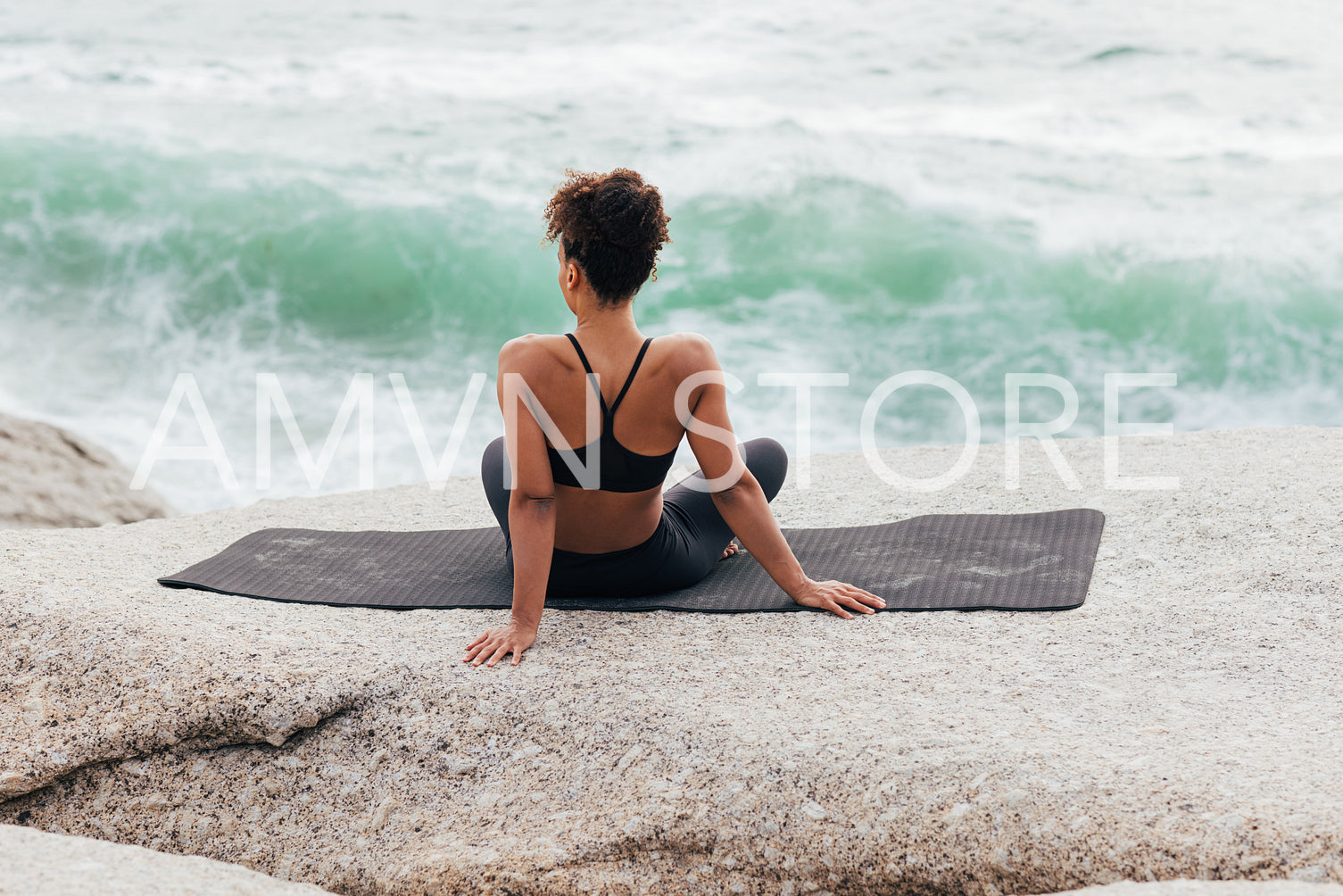 Rear view of young muscular woman sitting on yoga mat and looking on waves