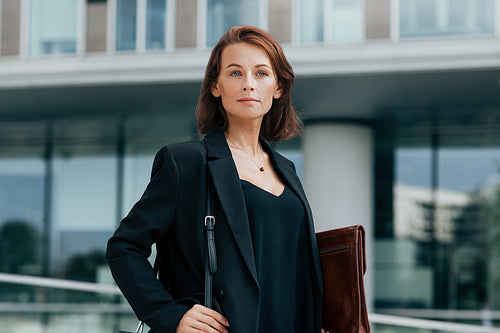 Confident middle-aged business woman with bag and leather folder standing against office building