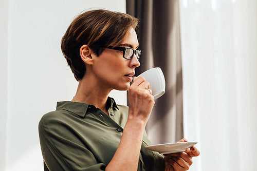 Side view of mid adult female in eyeglasses having coffee in hotel room standing at window