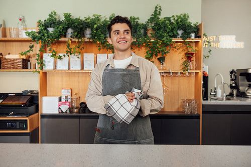 Smiling waiter in apron wiping hands with a towel while standing at the counter. Male barista in an apron with a towel.