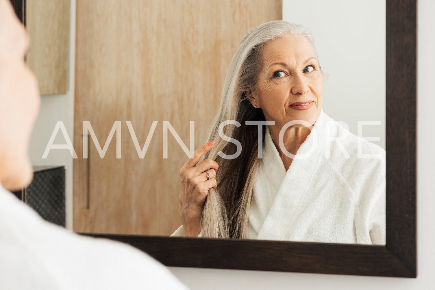 Senior woman examine her long grey hair while standing in front of a bathroom mirror