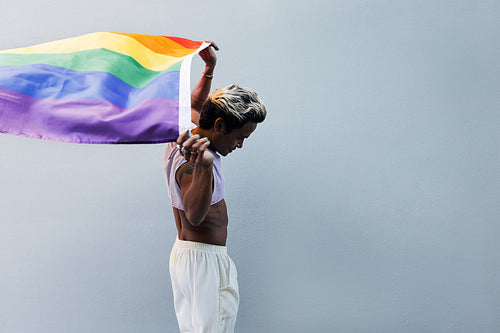 Young stylish man walking at grey wall raising hands with LGBT flag