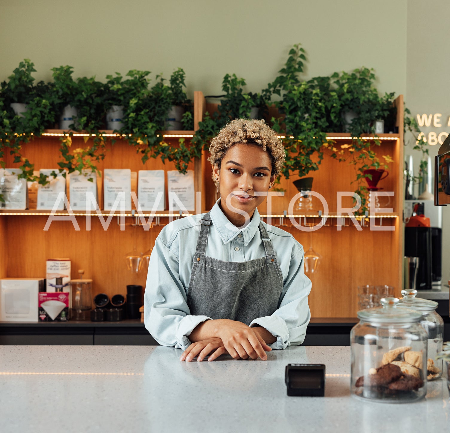 Young woman barista in an apron looking at the camera while standing at a counter in a small cafe