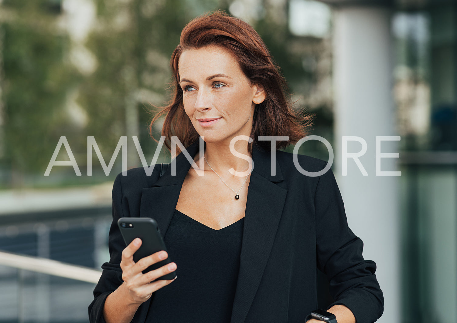 Confident female with ginger hair holding a smartphone and looking away. Middle-aged corporate person standing outdoors.