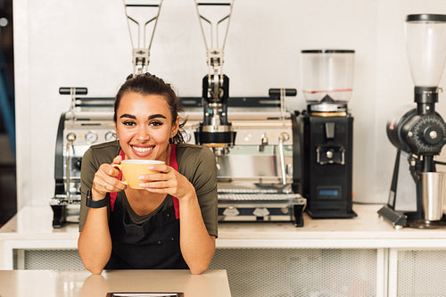 Smiling woman in apron leaning on a table while drinking coffee. Portrait of a waitress standing in front of the coffee machine.