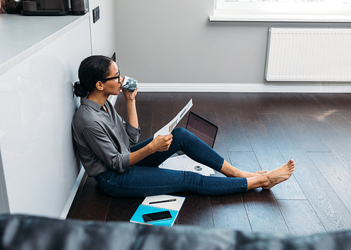 Young woman sitting on floor in living room and drinking coffee. Self employed female looking on document at home.