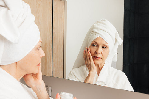 Senior woman with wrapped towel on her head applying cream on a face. Aged female doing morning routine in front of a bathroom mirror.